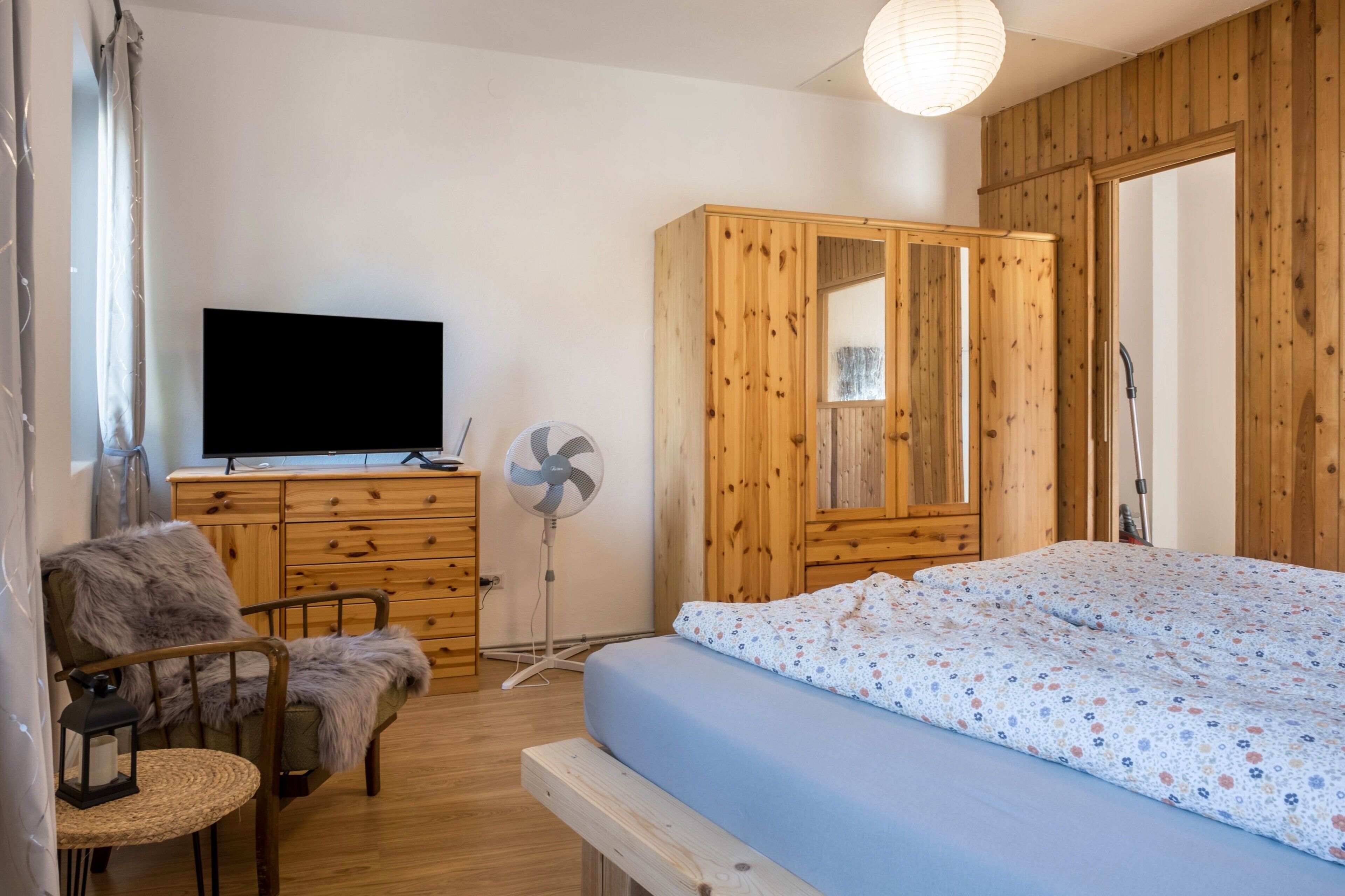 A room with a bed, a wooden wardrobe, a television, a fan, and a chair.