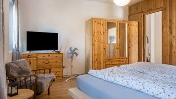 A room with a bed, a wooden wardrobe, a television, a fan, and a chair.