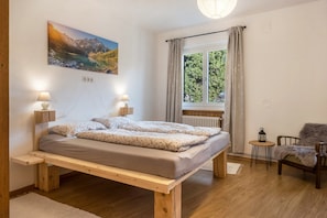 A bedroom with a wooden bed, bedside lamps, a view of greenery through a window, and a chair.