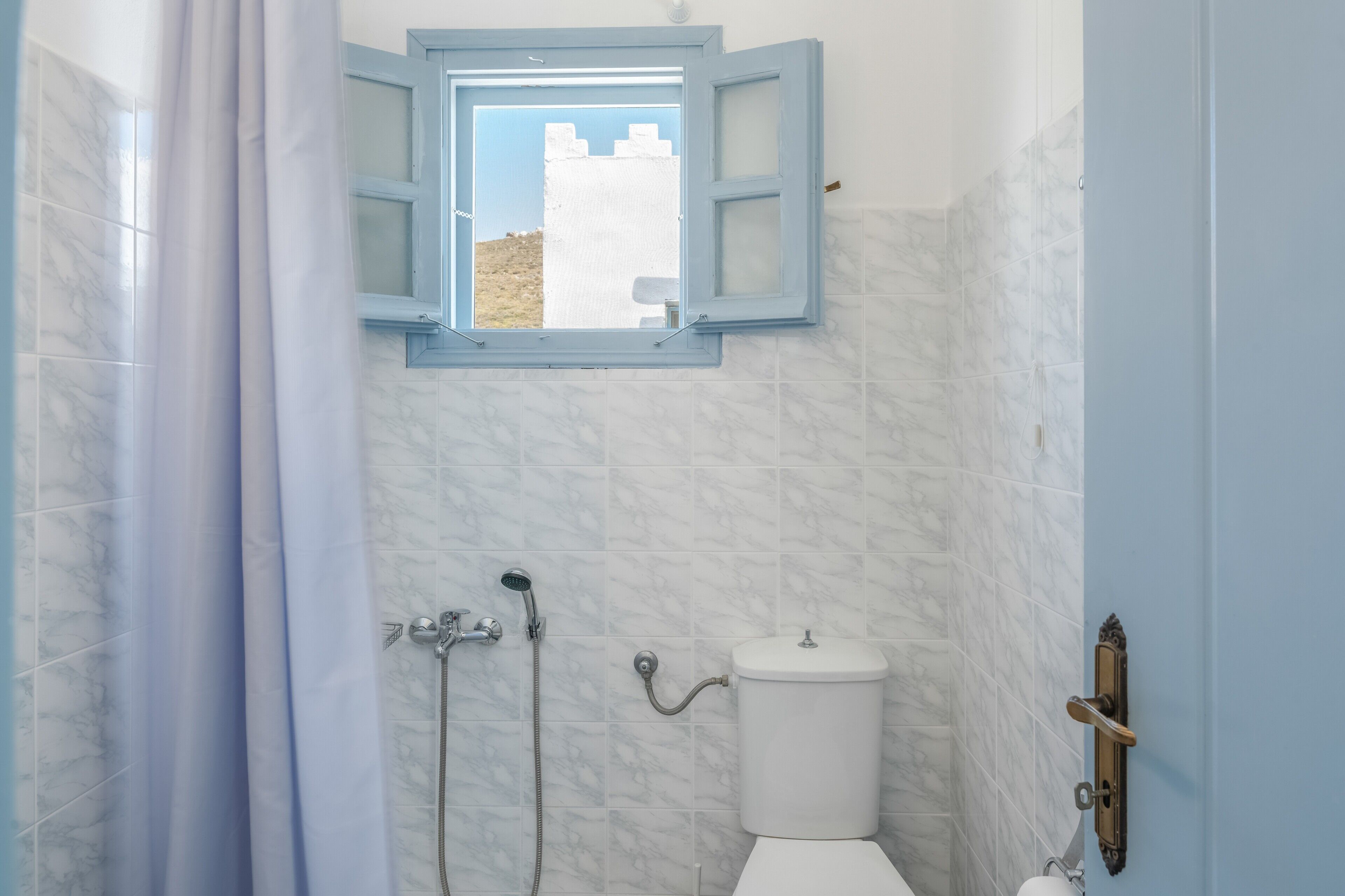 A bathroom with a toilet, shower, and a window with a view of a white building.