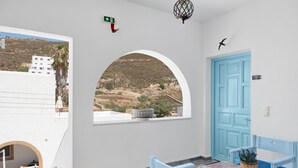 Une pièce avec une porte bleue, une fenêtre donnant sur une colline, une petite table avec une plante et un banc.