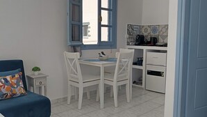 Et kompakt værelse med en seng, et spisebord med stole, et lille tekøkken med kaffemaskine og et vindue med blå skodder.