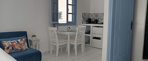 A compact room with a bed, a dining table with chairs, a small kitchen area with a coffee maker, and a window with blue shutters.