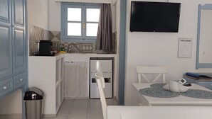 A compact kitchen with a refrigerator, a sink, and a window with curtains. There's a white table with a decorative mat and a chair. A TV is mounted on the wall, and a welcome sign is visible.