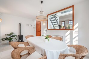 A dining area with a round table, wicker chairs, a white tablecloth, a potted orchid, a hanging light fixture, and a large mirror reflecting a staircase.