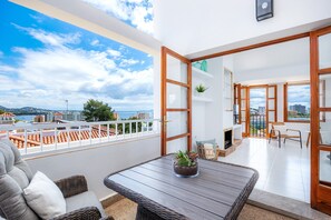 A balcony with a view of the city, a table with a plant, wicker furniture, and a door leading to an interior space.