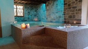 Un espacio tipo spa con paredes de piedra, una bañera caliente con azulejos, velas y vasos sobre una superficie texturizada.