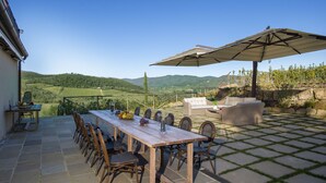 Un patio al aire libre con una mesa y sillas de madera, un paraguas grande y vistas a colinas ondulantes.