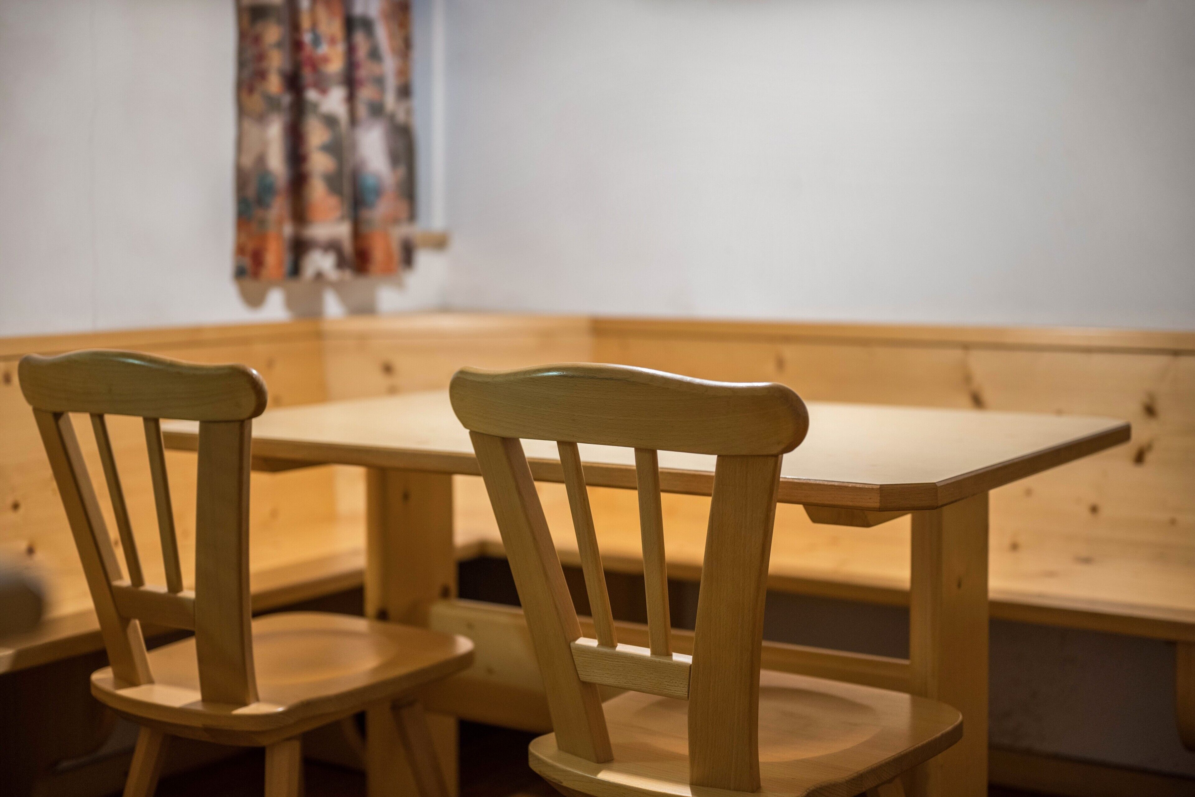A wooden table and two chairs in a room with a plain wall and a patterned curtain.