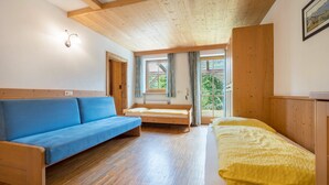 Una habitación con techo de madera, sofá azul, cama con manta amarilla y ventana con cortinas.