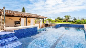 En swimmingpool med blå fliser, en pergola og en terrasse med grill.