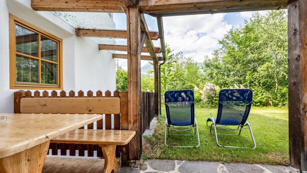 A wooden bench and table outside a building with a stone floor, a wooden fence, and a grassy area with two blue chairs.