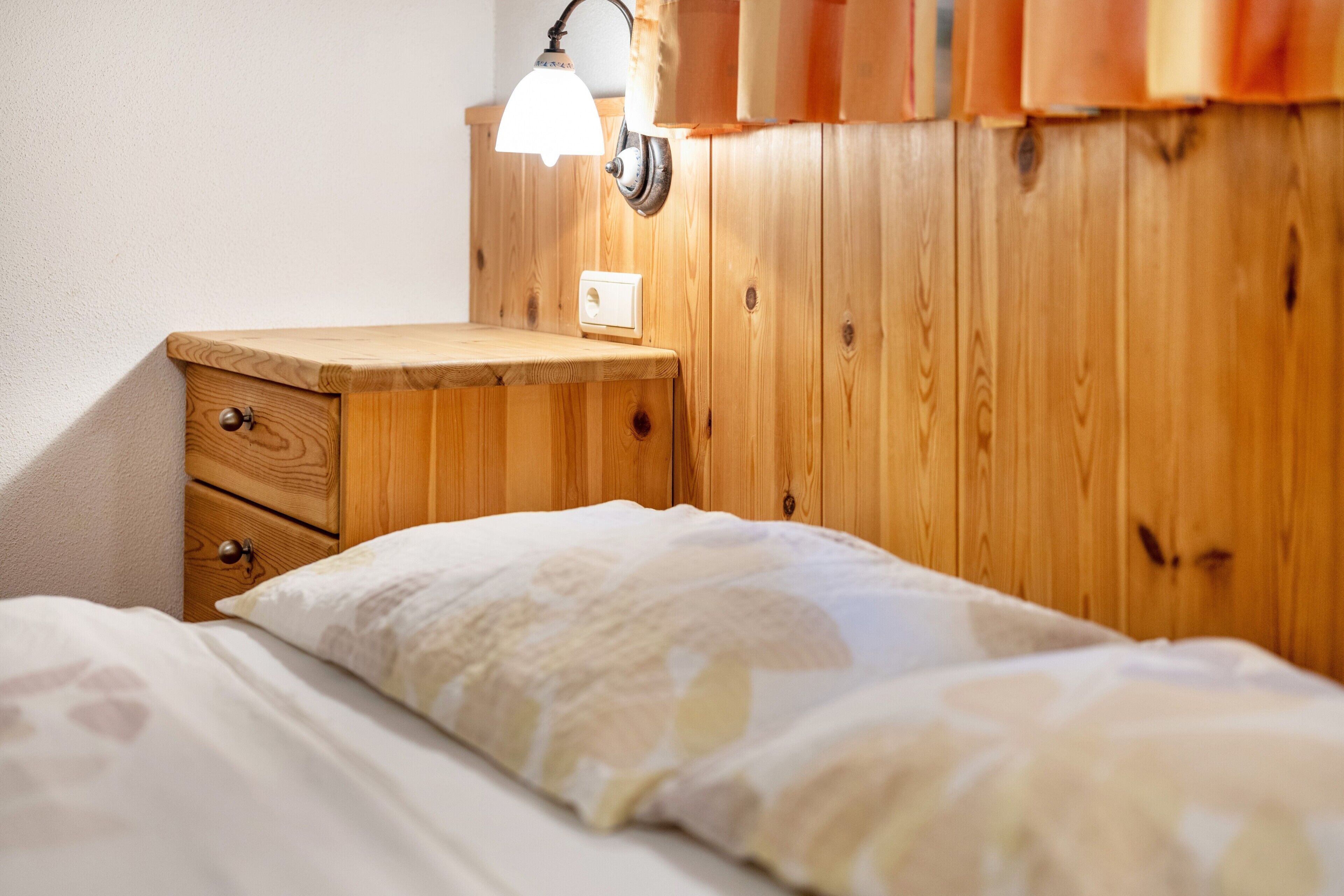 Ein Nachttisch aus Holz mit Schubladen, eine Lampe und ein Bett mit einem gemusterten Kissen.
