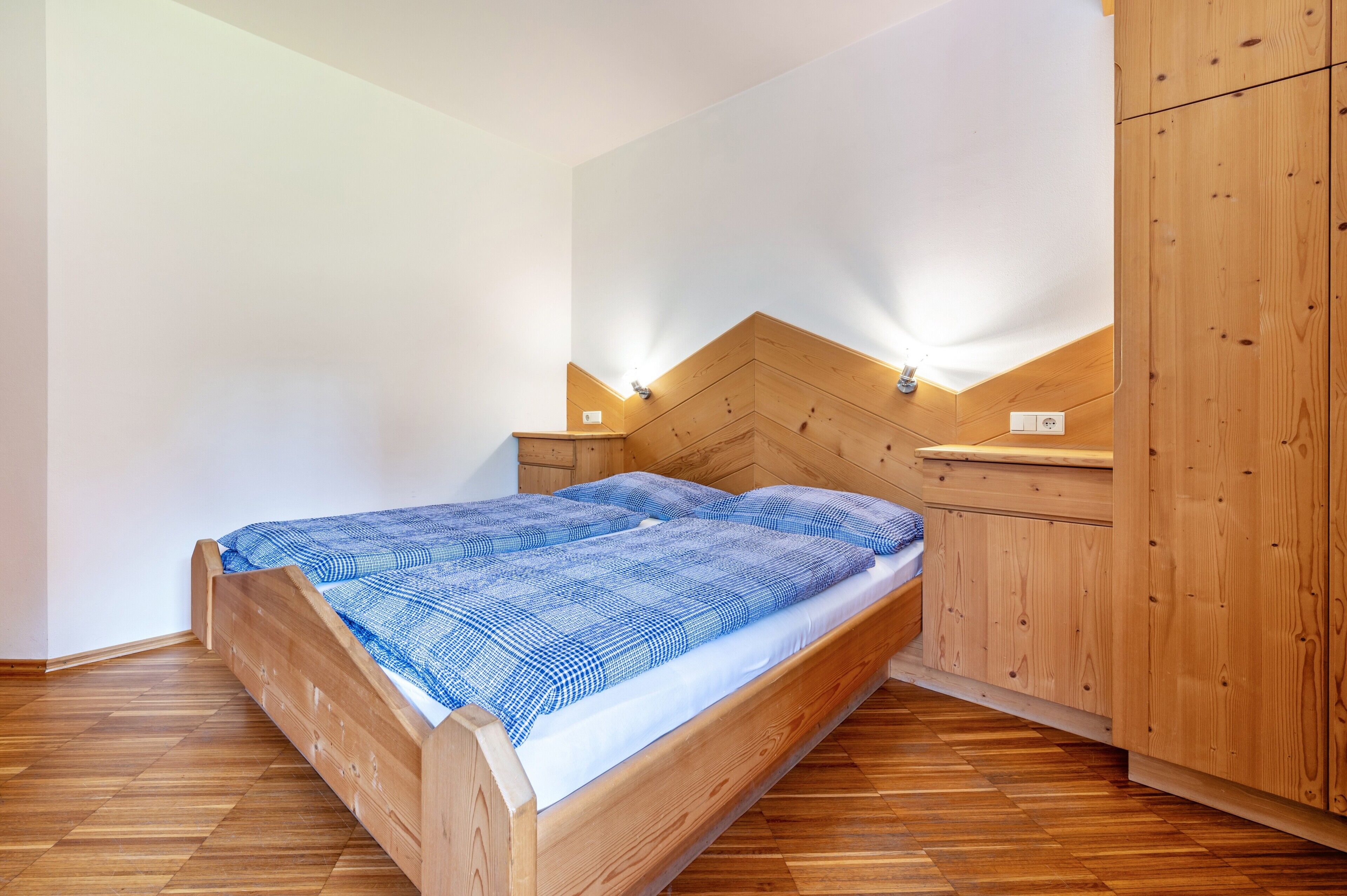A single bed with a wooden headboard, a nightstand, and a wooden wardrobe in a room with wooden flooring and white walls.
