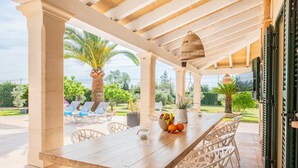 An outdoor patio with a wooden table, white chairs, a hanging light fixture, and a view of a garden with palm trees. - Holiday Home 'Villa Sun Club Home' with Private Pool, Wi-Fi and Air Conditioning (Llucmajor)