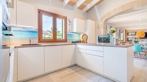 A modern kitchen with white cabinetry, a wooden ceiling, and a view of the beach through a window. - Holiday Home 'Villa Sun Club Home' with Private Pool, Wi-Fi and Air Conditioning (Llucmajor)