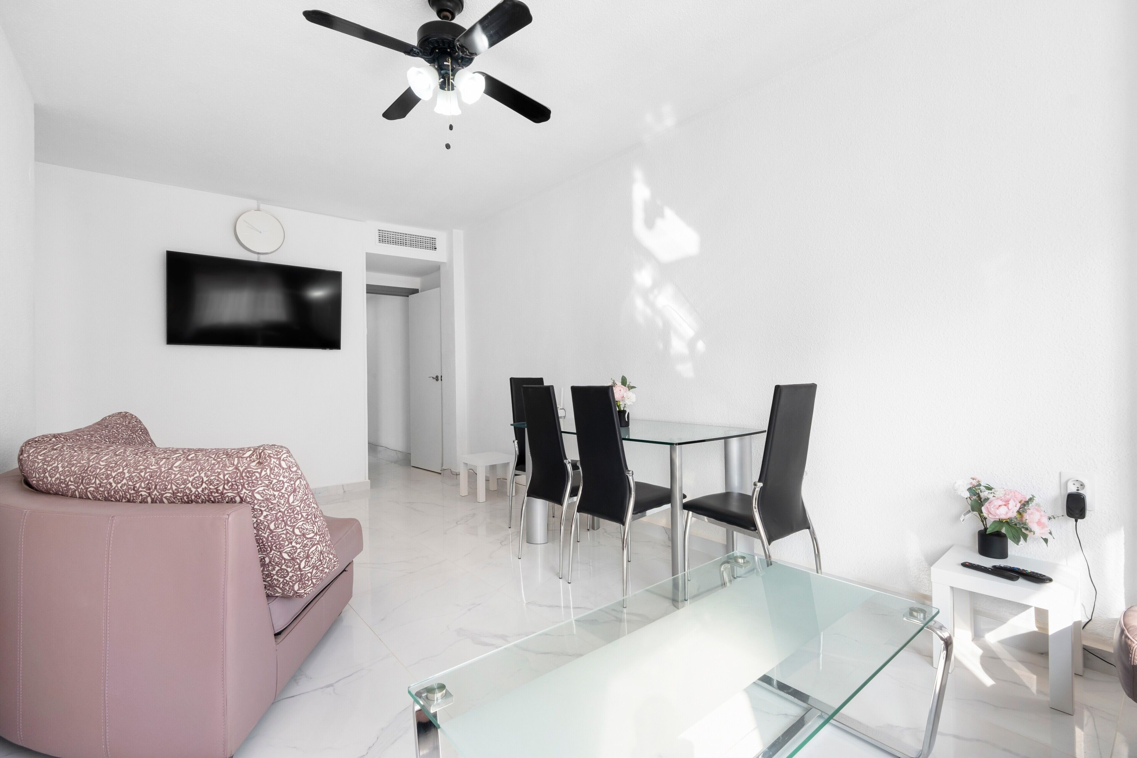 Una sala de estar moderna con una mesa de centro de vidrio, un sofá rosa, sillas negras, un ventilador de techo y un televisor montado en la pared.