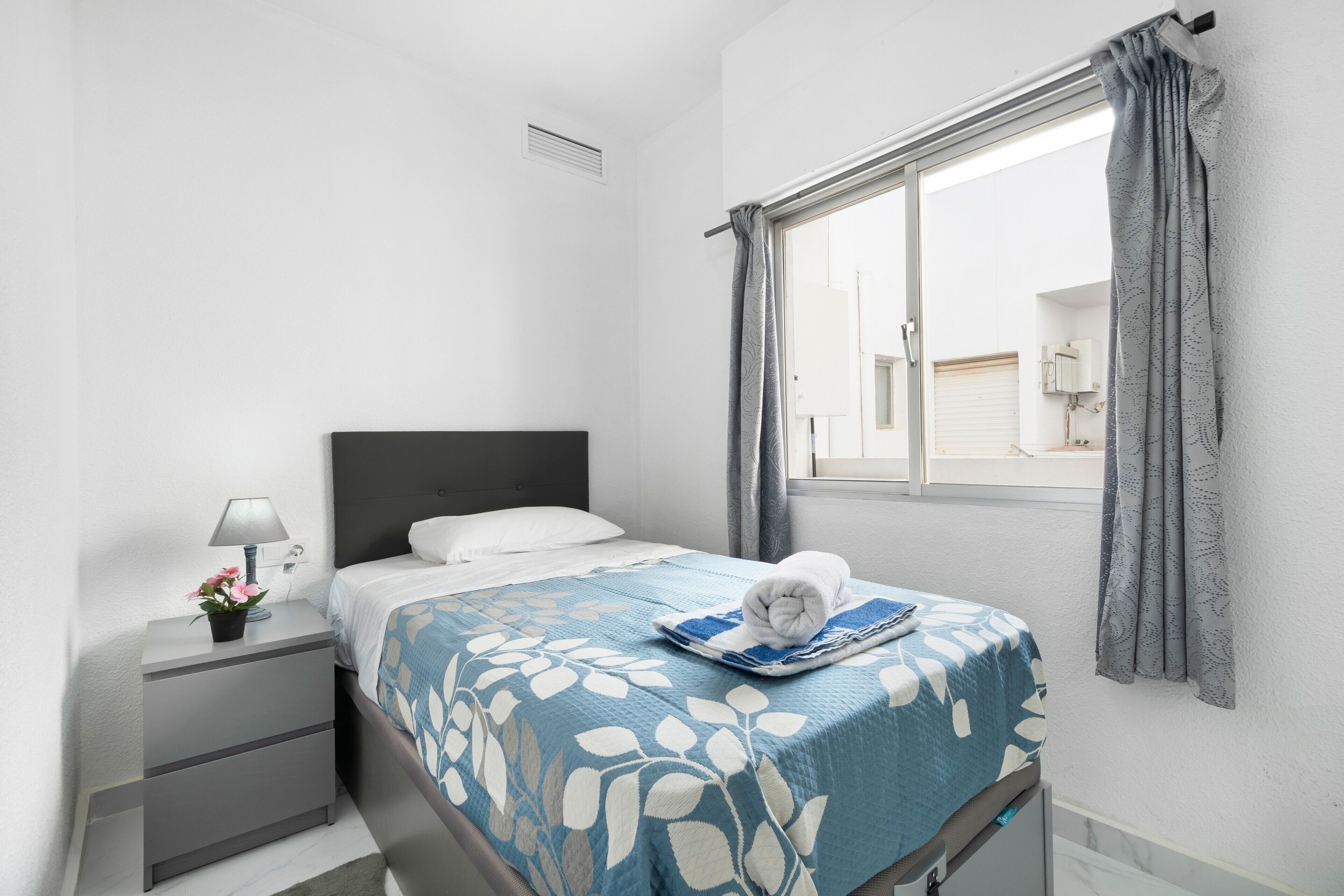 A bedroom with a bed, a nightstand, a lamp, a window with curtains, and a view of the outside.