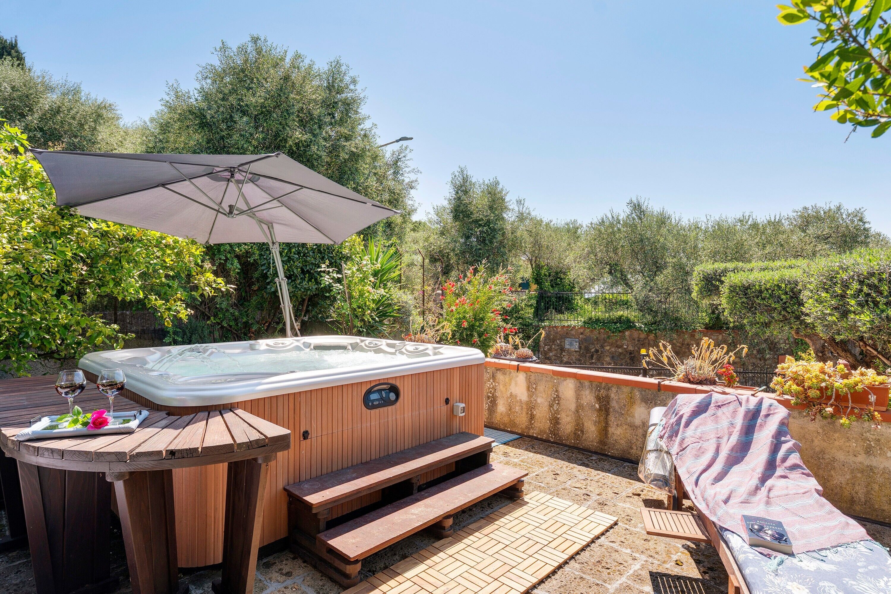 Uma banheira de hidromassagem com degraus, uma mesa de madeira com duas taças de vinho e um guarda-sol.