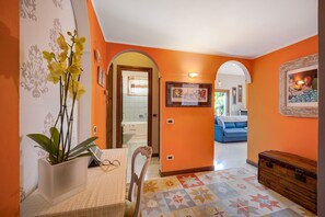 Um quarto com uma cama, uma cadeira, uma mesa com um vaso de flores, um quadro na parede e um arco que dá para outro quarto.