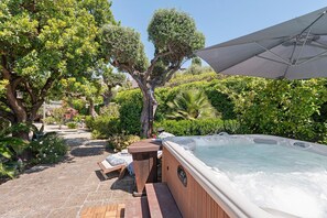 Un jacuzzi en un jardín con terraza de madera, sillas de descanso y una sombrilla.