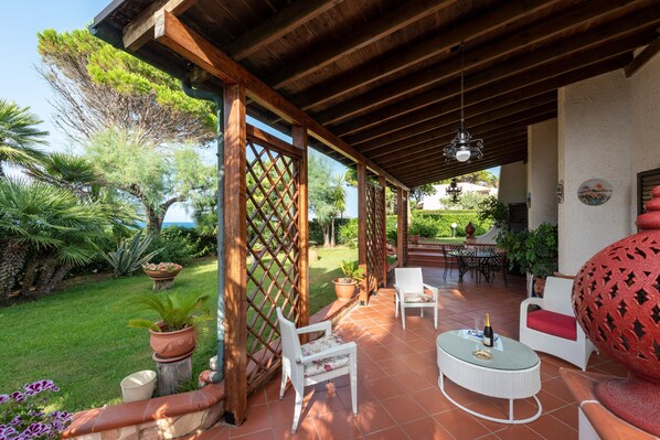 An outdoor patio with wooden beams, white wicker furniture, and potted plants.