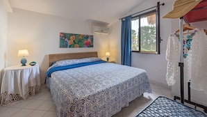 Ein Schlafzimmer mit Bett, Nachttisch, Lampe, einem Gemälde, einer Klimaanlage, einem Fenster mit Vorhängen und einem Hut auf einem Ständer.