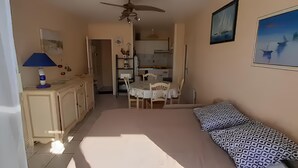 Une pièce comprenant un lit, un bureau, une chaise, une lampe, un ventilateur de plafond, un tableau et, au fond, un coin cuisine.