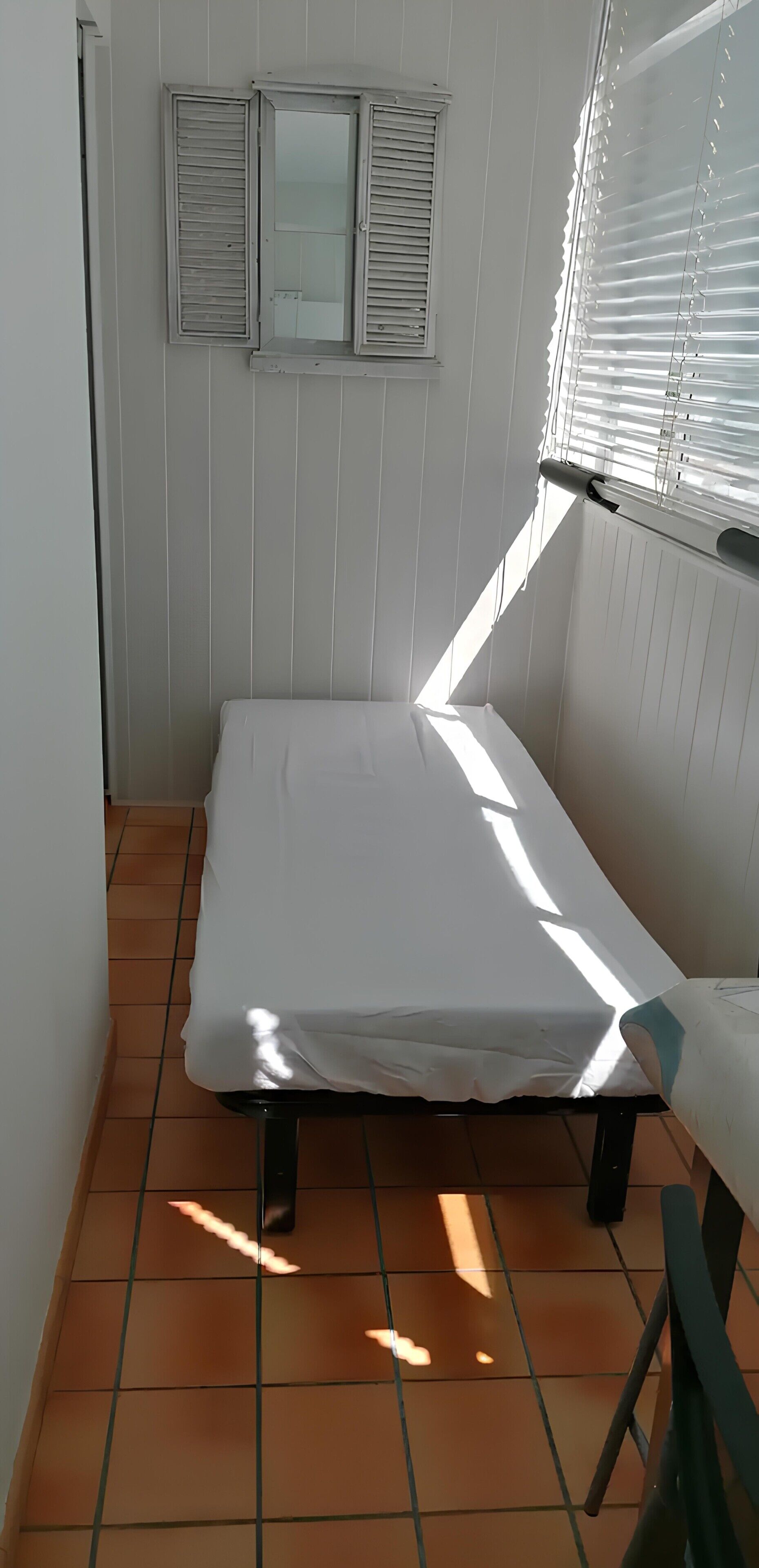 A small, single-bed room with a tiled floor, a window with blinds, and a radiator.