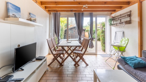Ein modernes Wohnzimmer mit Fernseher, einem Holzespeisetisch und Stühlen, einer Couch und Blick ins Freie.