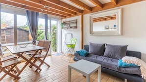 Ein gemütlicher Wohnbereich mit einer Couch, einem Couchtisch und einem Esstisch mit Stühlen. Ein großes Fenster mit Vorhängen, ein Spiegel an der Wand und eine hängende Pflanze sind ebenfalls vorhanden.