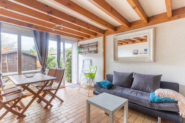 A cozy living area with a sofa, a coffee table, and a dining table with chairs. There is a large window with curtains, a mirror on the wall, and a hanging plant.