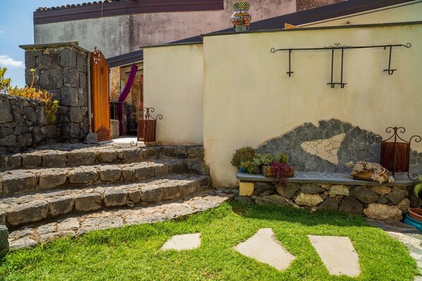 Un escalier en pierre menant à une porte ornée d’un rideau violet, un mur de pierre agrémenté d’une arche décorative et un petit jardin avec des plantes en pot.