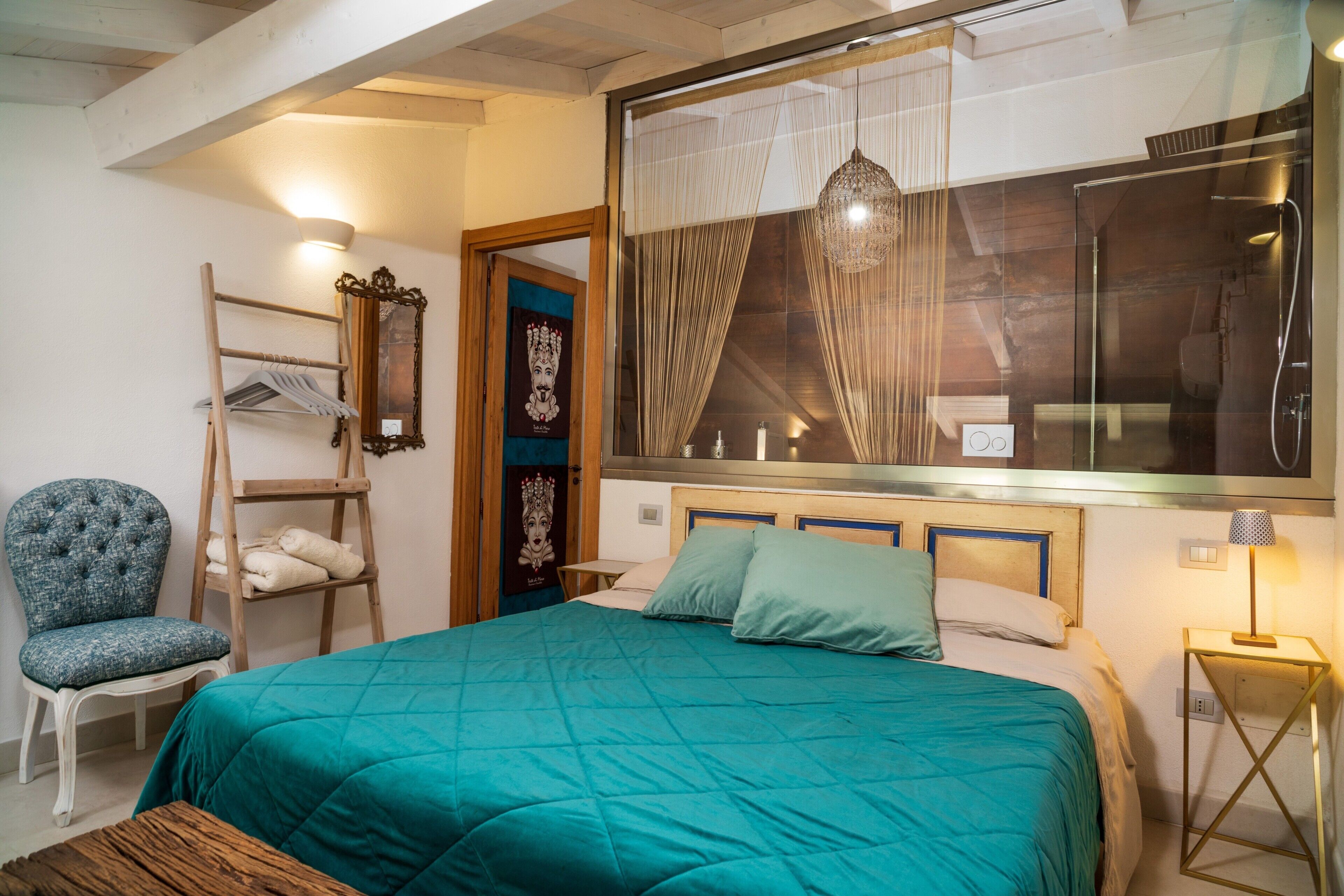 Un dormitorio con cama, una silla, estante, espejo y ventana con cortinas.