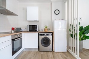 A compact kitchen with a microwave, oven, washing machine, and a clock on the wall.
