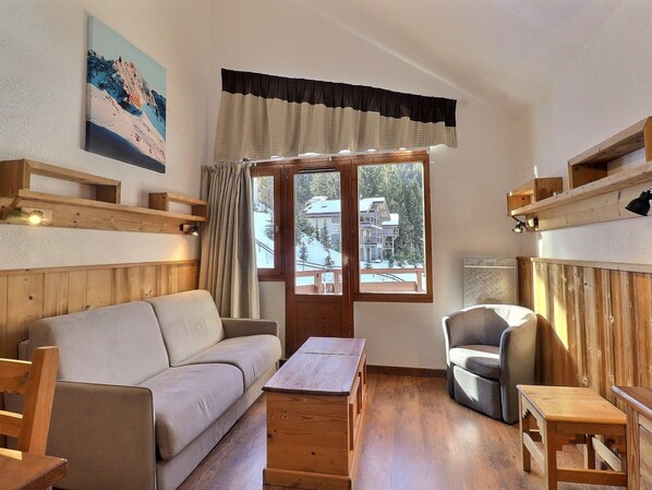 Une pièce chaleureuse avec un plancher en bois, un canapé, un fauteuil, une table et une fenêtre donnant sur un immeuble et des arbres.