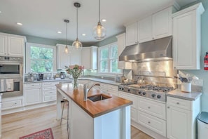 A modern kitchen with white cabinetry, a central island with a sink, stainless steel appliances, pendant lighting, and a view of greenery outside. - Spacious Island Escape Near Beach and Marina (Isle of Palms)