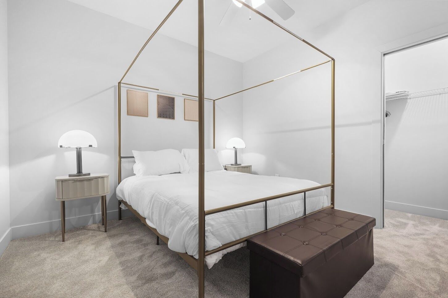 A four-poster bed with a bench, two bedside tables, and a ceiling fan in a minimalist room with white walls and carpet.