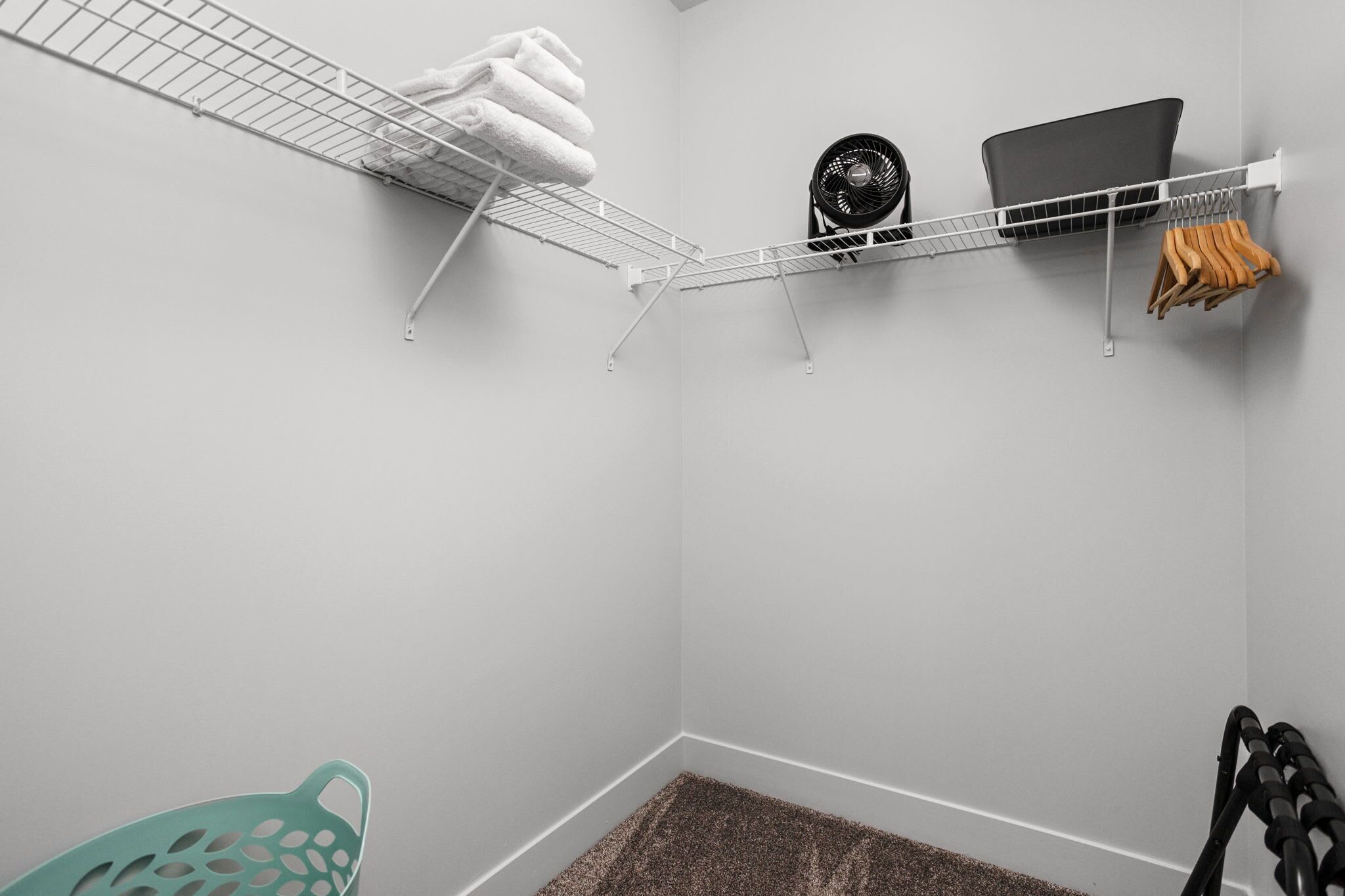 Esquina de una habitación con pared blanca, estante metálico con toallas, un ventilador y una computadora portátil.