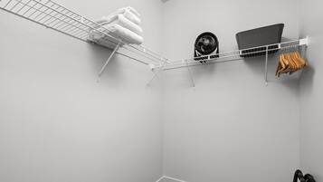 Esquina de una habitación con pared blanca, estante metálico con toallas, un ventilador y una computadora portátil.
