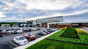 Un centro comercial moderno con varias tiendas, un estacionamiento lleno de coches y un área ajardinada con césped y plantas.