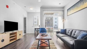 A modern living room with a flat-screen TV, a wooden coffee table, a black leather sofa, and a dining area with a blue chair. - CozySuites at Notch (Indianapolis)