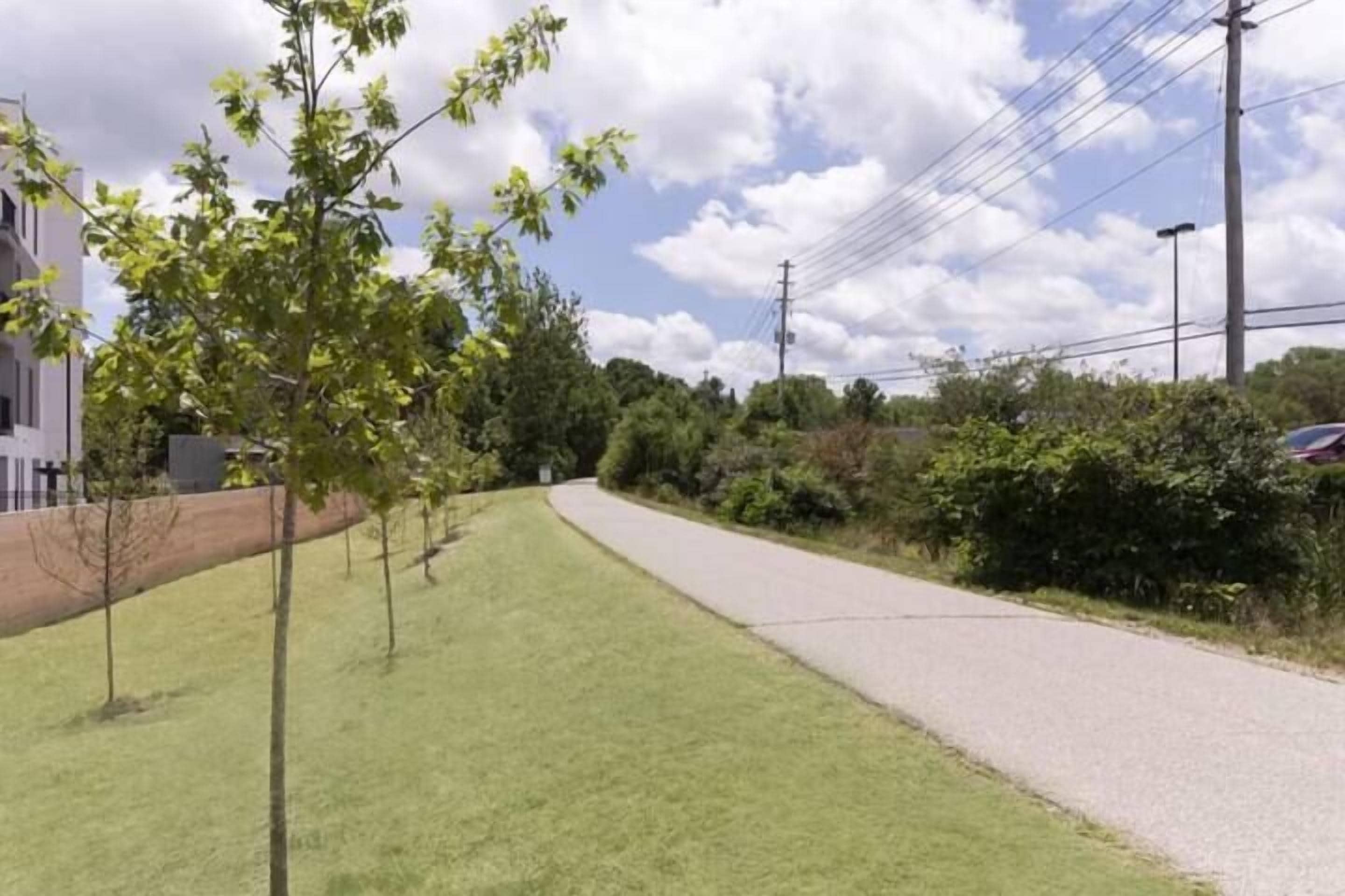 A paved pathway alongside a grassy area with trees and a building in the background.