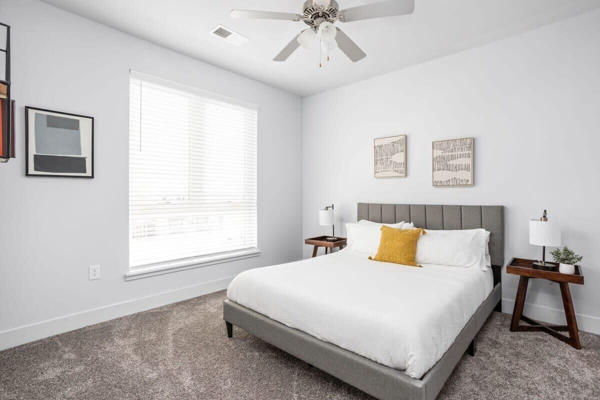 A bedroom with a large bed, a ceiling fan, a window with blinds, and two bedside tables with lamps.