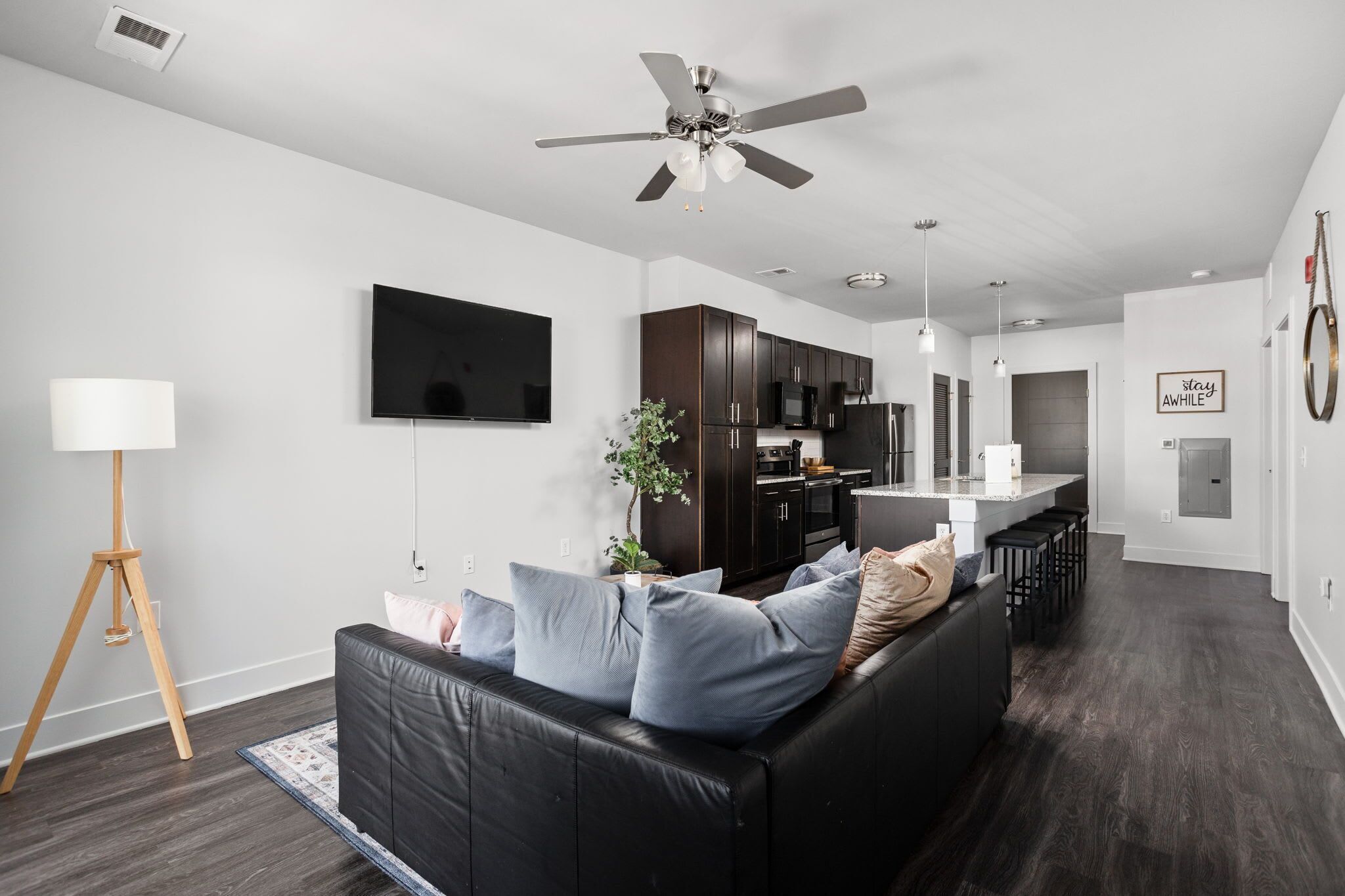 A modern living room with a black leather sofa, a flat-screen TV, a ceiling fan, and a kitchen area in the background.