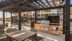 An outdoor patio with wooden tables and chairs, a grill, and a pergola structure. - CozySuites at Notch (Indianapolis)