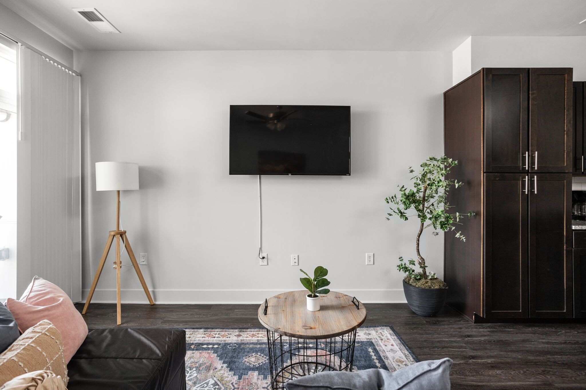 Una sala de estar moderna con un televisor de pantalla plana montado en la pared, una lámpara de pie, una mesa redonda de centro con una planta, un mueble de madera oscura y un amplio ventanal con cortinas.