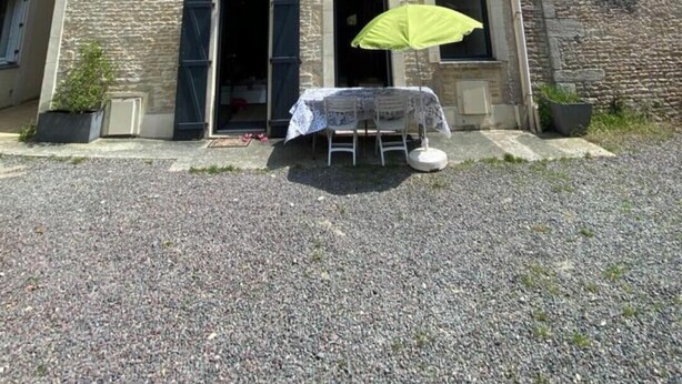 Een tweelaags stenen gebouw met een grindpad, een tafel met een gele parasol, en een kleine tuin.