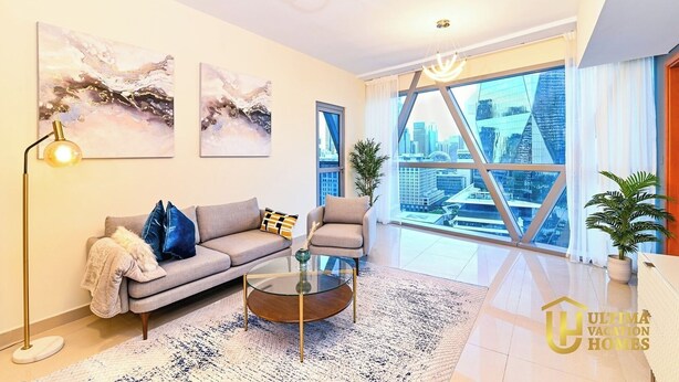 Una sala de estar moderna con un sofá, un sillón y una mesa de centro, ventanas amplias con vistas a la ciudad y arte abstracto en la pared.