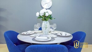 Una mesa de comedor preparada para cuatro personas, con un jarrón de flores blancas, un reloj de pared y el logotipo de Ultima Vacation Homes.